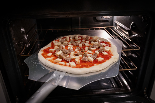 A pizza steel inserts a homemade sausage pizza into an oven onto a preheated pizza steel.