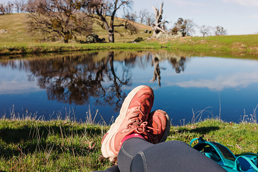 Person in hiking shoes resting on the grass near the small lake after the hike in spring