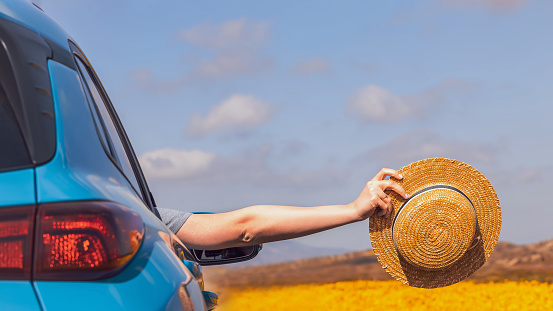 Female hand holding a straw hat in her hand stretched out from the passenger seat window of the car