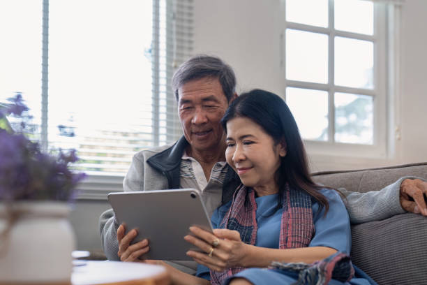 istock Happy elderly asian couple using tablet sit on sofa doing ecommerce shopping online on website and buying insurance browsing at home 2067090632