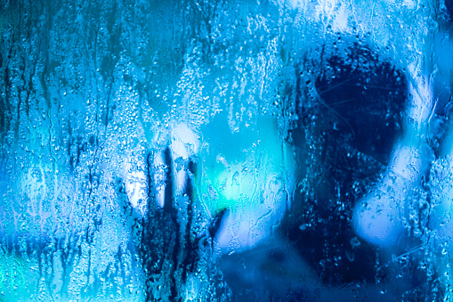 A hand is shown through a window with raindrops on the glass. Scene is somber and melancholic, as the hand appears to be reaching out for something that is not visible