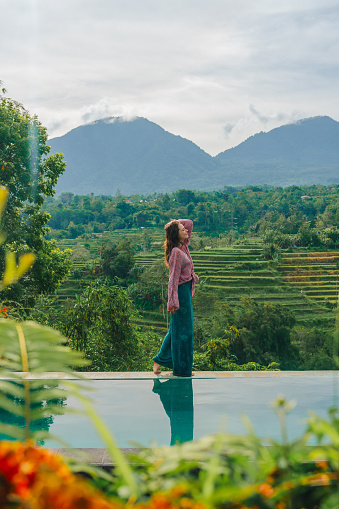 Elegant woman standing on the edge of swimming pool overlooking rice fields and mountains on Bali
