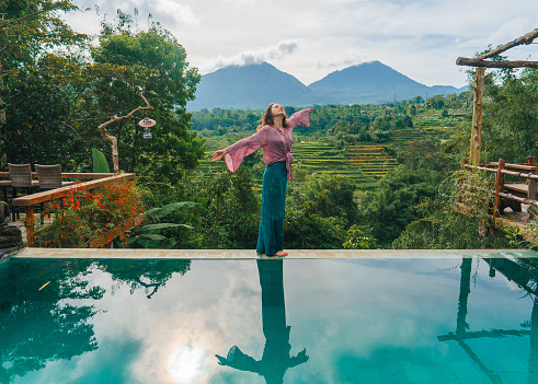 Elegant woman standing on the edge of swimming pool overlooking rice fields and mountains on Bali