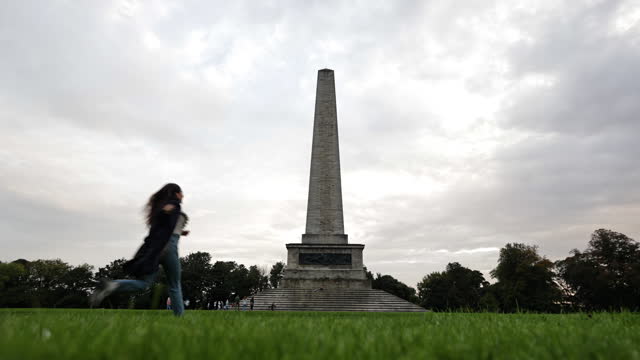 Young woman running happily drops her clothes on the ground, Wellington Monument, Wellington Monument in Phoenix Park in Dublin Ireland, famous tourist attraction in Ireland, landscaped park in Europe, famous city parks in Europe