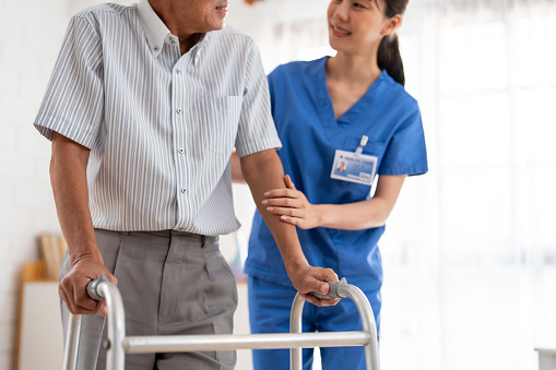 Close up of senior man patient doing physical therapy with caregiver. Attractive specialist carer women help and support older mature male practice walking slowly with walker at nursing home care.