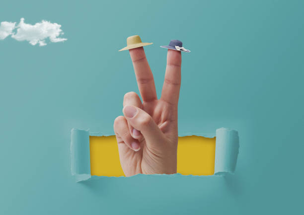 Female hand making a V sign and wearing tiny beach hats on two fingers, the hand is coming out from a hole in torn paper, summer vacations concept