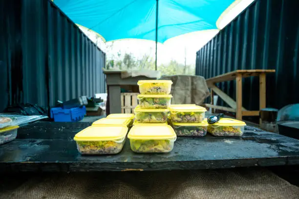 prepared meal containers lined up on a rustic workbench at an outdoor campsite with a blue tarp overhead, showcasing organized food prep for camping