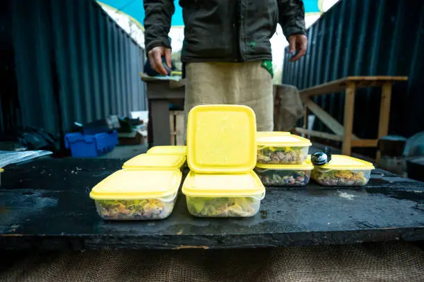prepared meal containers lined up on a rustic workbench at an outdoor campsite with a blue tarp overhead, showcasing organized food prep for camping