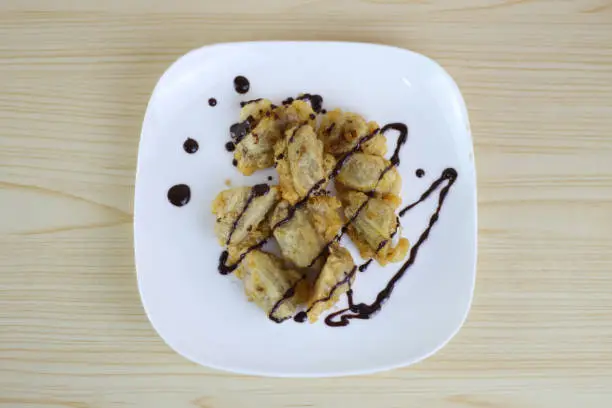 Photo of Pisang Goreng Keju or Fried Banana topped chocolate with on white plate on the wooden table.