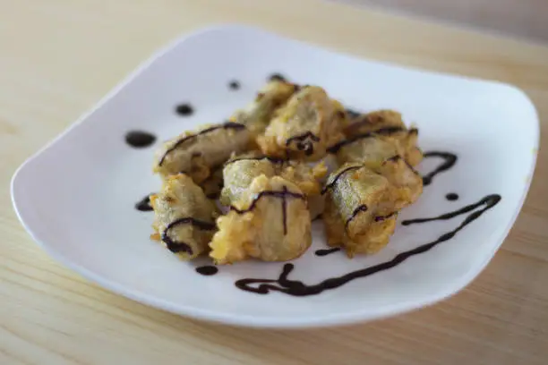 Photo of Pisang Goreng Keju or Fried Banana topped chocolate with on white plate on the wooden table.