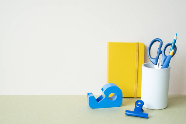 Yellow notebook, blue colored pencil, pen, scissors, clip, tape dispenser on desk. white ivory background. workspace office stationery