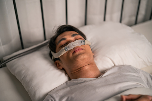 Asian man wearing cpap machine sleeping smoothly all night in bedroom. Attractive handsome healthy male feel happy and relax while lying down on bed, using sleep apnea oxygen mask therapy equipment.