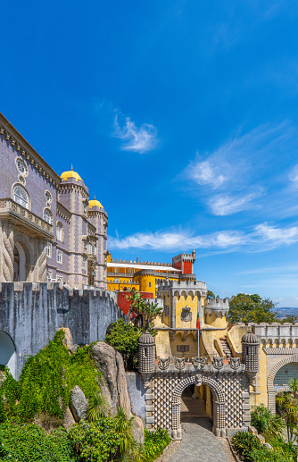 Exterior and wide view of the Pena Palace of yellow, red and mosaic colors with the path up to the arched entrance with bas-reliefs of the Palace, under a sunny blue sky. Sintra. Portugal.