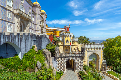 Exterior and wide view of the colorful Pena Palace with the path up to the arched entrance decorated with bas-reliefs, under a sunny blue sky. Sintra. Portugal.