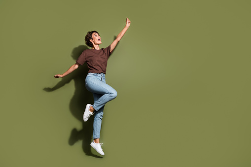 Full size photo of ecstatic woman dressed brown t-shirt pants flying catching object look empty space isolated on khaki color background.