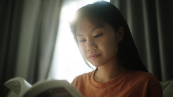 Asian teenage girl reading a book on sofa at home.