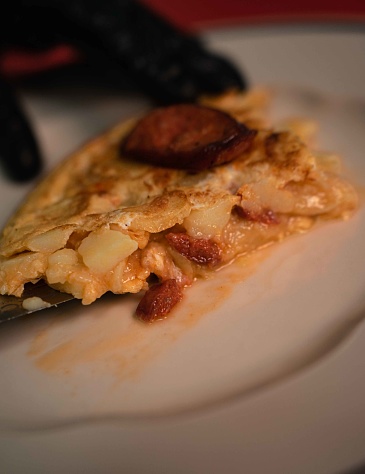 A tempting view of a Spanish potato omelette adorned with slices of chistorra sausage, a traditional delicacy from the Basque country. The golden hue of the omelette contrasts beautifully with the rich, reddish tones of the chistorra, creating an appetizing visual feast. This iconic dish represents the heart and soul of Spanish gastronomy, inviting you to indulge in its savory flavors and cultural heritage