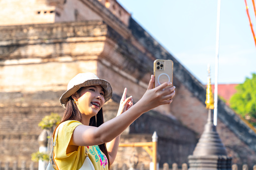 Happy Asian woman tourist using mobile phone taking selfie or vlogging during travel old Buddhist temple in Chiang Mai. Attractive girl enjoy outdoor lifestyle travel Thailand on summer holiday vacation.