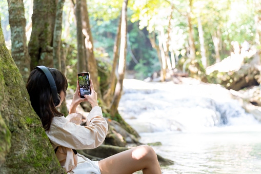 Young Asian woman travel nature on summer holiday vacation. Generation z girl enjoy and fun outdoor lifestyle using mobile phone taking selfie or vlogging with beautiful nature at forest mountain.