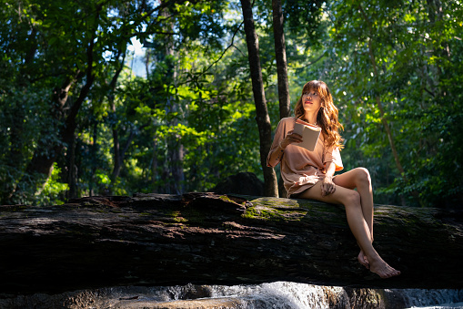 Happy young Asian woman enjoy and fun outdoor lifestyle travel nature forest mountain on summer holiday vacation. Attractive girl sitting on tree trunk and writing journey diary at nature waterfall.