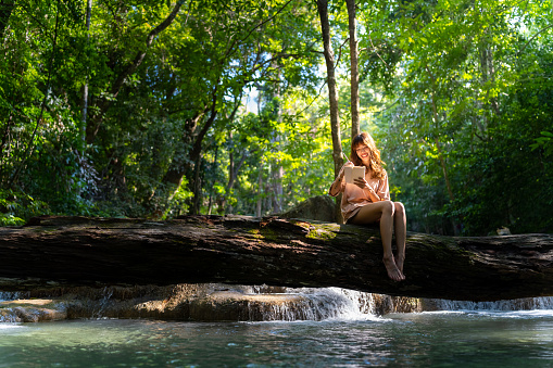 Happy young Asian woman enjoy and fun outdoor lifestyle travel nature forest mountain on summer holiday vacation. Attractive girl sitting on tree trunk and writing journey diary at nature waterfall.