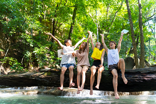 Group of Young Asian man and woman enjoy and fun outdoor lifestyle travel nature tropical forest on summer holiday vacation. Happy generation z people friends sitting on tree trunk and playing water together at waterfall lagoon.