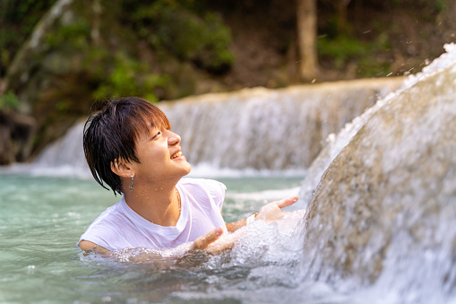 Young Asian man travel nature tropical forest on summer holiday vacation. Happy generation z guy relax and enjoy outdoor lifestyle swimming and relaxing in water at waterfall lagoon.