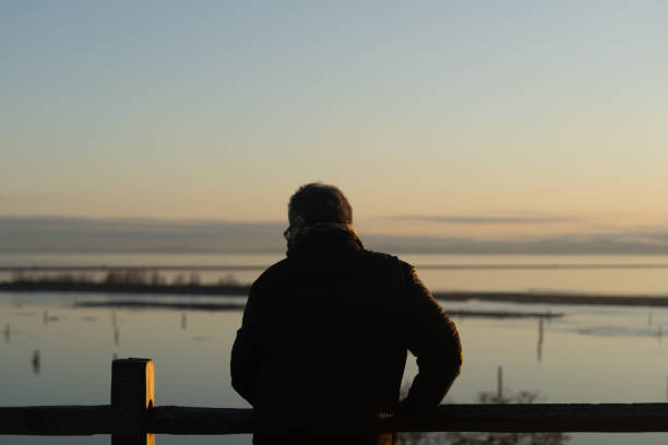 In this photograph, an individual in a black jacket stands quietly in front of a railing, with their back to the camera, gazing out at the calm sea during dusk. Silhouettes of small boats scattered across the water complement the gentle orange hues of the sky, creating an atmosphere of depth and tranquility. The scene captures a moment of personal reflection alongside the grandeur of the natural world, inviting contemplation of life's peaceful moments.