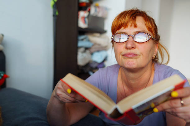 Red-haired woman with glasses emotionally reads book, expression emotions and passion on face, in home interior