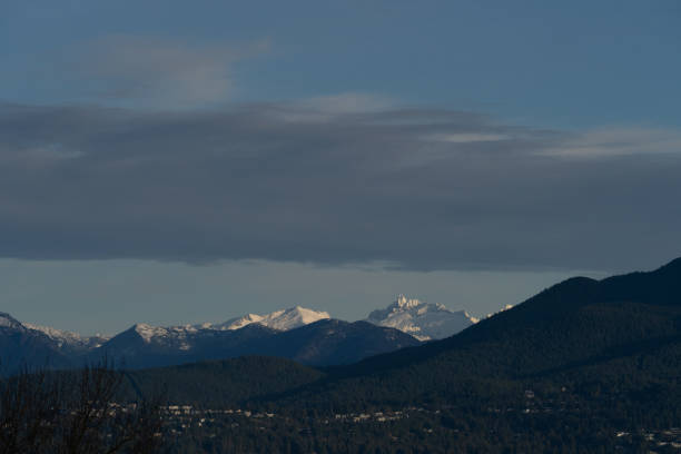 Captured from a vantage point at the University of British Columbia (UBC), the photograph showcases the beautiful rolling hills of West Vancouver adorned with fresh snow. The blue-grey clouds stretch across the sky, offering a splendid canvas for the sunlit peaks. This scene depicts the tranquility and grandeur of Vancouver's winter landscape and the harmonious coexistence of the city with the natural world.