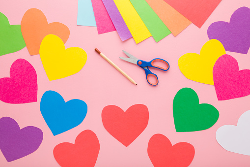 Colorful heart shapes, scissors, pencil and application paper on light pink table background. Pastel color. Closeup. Making decoration elements. Top down view.