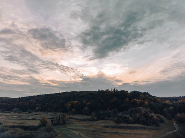 Aerial moody autumn rural scenery with cloudy grey sky, dirt road in fields and forest