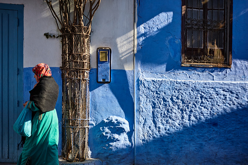 Blue city street with Moroccan woman walking in colourful traditional clothing side view in Chefchaouen, Morocco, North Africa.