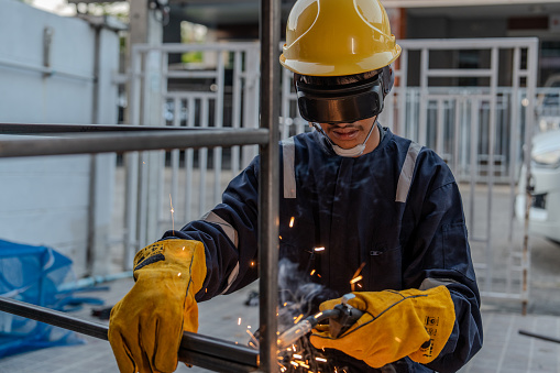 Asian worker wearing goggles, gloves, hat and safety suit uses a welding machine. Sparks from welding wire and steel create sparks.