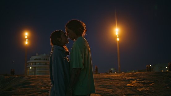 Loving teenagers looking eyes to eyes at night beach. Romantic pair dating at evening zoom on. Smiling man whispering tender words to lovely girlfriend. Affectionate couple enjoying moment together
