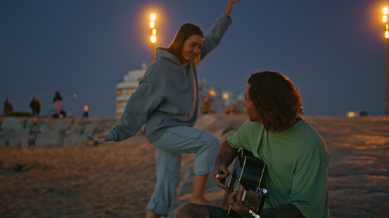 Young woman embracing boyfriend at evening sandy beach. Handsome man playing guitar at twilight nature. Romantic sweethearts resting together at vacation. Happy lady listening song enjoying music