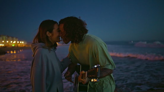 Enamoured man playing acoustic guitar at night beach closeup. Curly young guy serenading at musical instrument kissing girlfriend at evening. Relaxed carefree sweethearts spending free time in nature