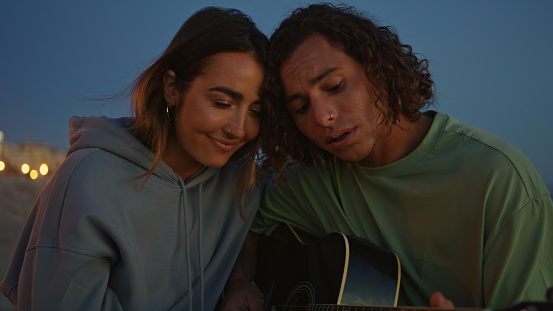 Singing man playing guitar at evening close up. Romantic young woman bonding to curly boyfriend listening music at dusk nature. Love couple enjoying each other dating at sandy beach. Happiness concept