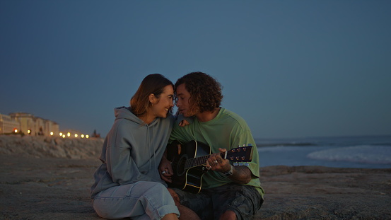 Romantic pair enjoying guitar music at evening sea nature. Curly man playing song serenading for girlfriend at twilight. Relaxed woman listening acoustic sounds on sand beach surface. Happiness love