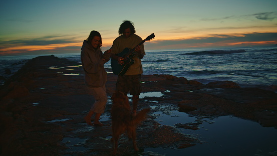 Carefree teenagers silhouettes relaxing morning seaside. Relaxed young man playing guitar at sunrise beach. Woman dancing at ocean horizon background. Unrecognizable love couple having fun with dog