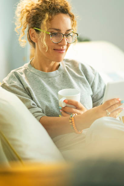 young woman reading ebook on electronic reader tablet sitting comfortably on the sofa at home in indoor leisure activity. people with ereader enjoying relax indoor on couch. happy female people inside - e reader cheerful indoors lifestyles fotografías e imágenes de stock