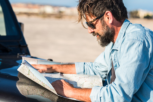 One traveler man looking paper guide map on car hood outside to find best roads and arrival destination. Transport and travel with vehicle. Driver lost in problem. Handsome tourist enjoy drive