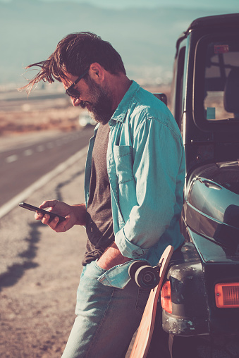 One man travel with car and standing outside the vehicle using online mobile phone app maps to choose roads and next destination with long scenic roads in background. Concept of transport technology