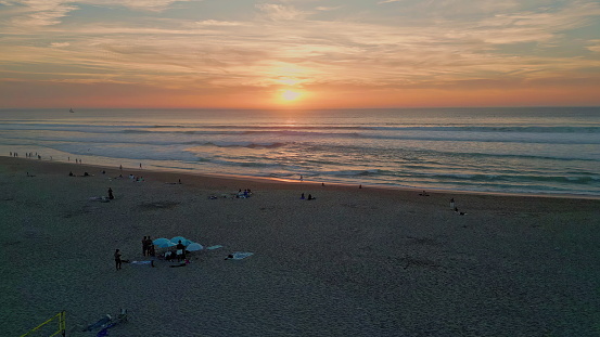 Orange ocean sunset shining on endless marine horizon summer evening. Diverse people resting on sandy beach at vacation drone view. Majestic golden sun setting down over foamy sea waves in slow motion