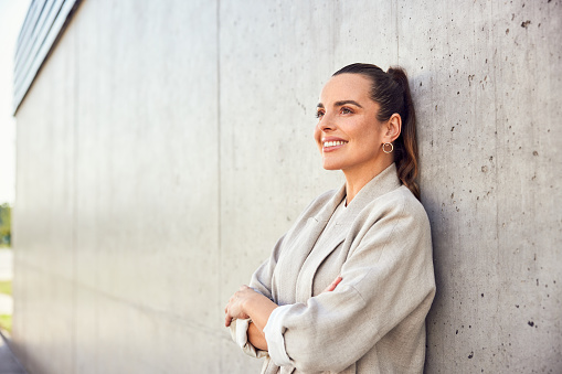 Adult successful businesswoman daydreaming looking up standing against concrete wall in urban scene