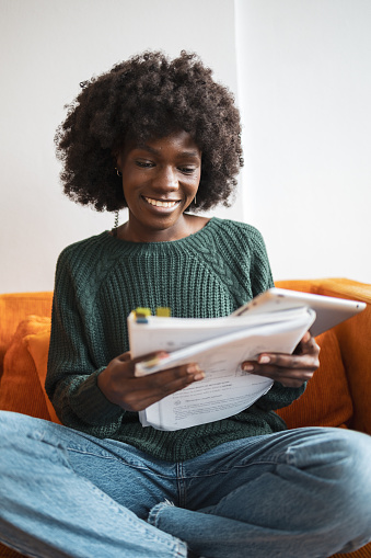 Smiling woman sitting at home and studying. She is holding the book and digital tablet