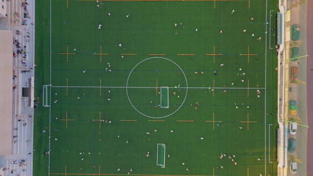 Aerial view green football field with training commands at summer evening. Top shot beautiful modern grass stadium with fans on grandstand. Small town playground for soccer game championship zoom out.