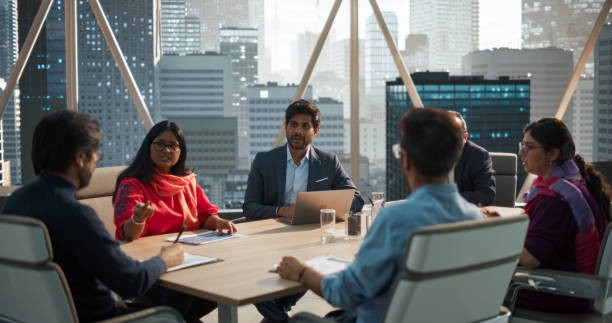 Diverse Team of Indian Managers Talk, Brainstorm, Use Laptop Computer and Printed Documents. Businessman Presenting Investment Strategy to Partners. Modern Multiethnic Office Conference Room Meeting