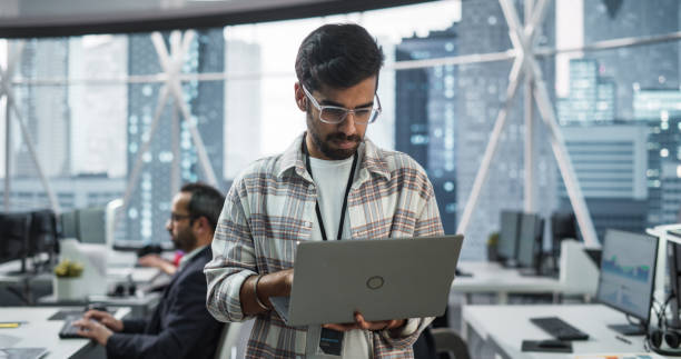 istock Professional Indian Specialist Using Laptop while Standing in an Office Near a Window with a Big City View. Successful Software Data Engineer Optimizing Corporate Database, Update Security Network 2068235636
