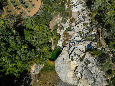 Drone view of a natural pool of crystal clear green water with a waterfall located between some rocks in the middle of a lush forest.
